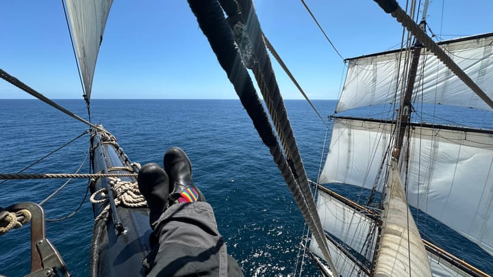 Person sitting on a square rigged mast with their legs stretching into the distance towards the horizons. Sails billow on the foremast ahead of them.